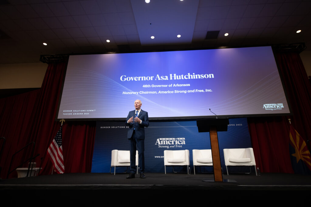 Former Governor Asa Hutchinson speaks in front of a screen bearing his name and the conference title, "America Strong and Free Summit"